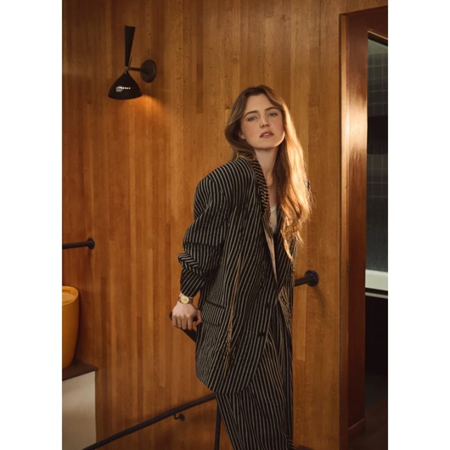 Woman with long brown hair poses in a striped pantsuit on wooden stairs next to a yellow vase and black sconce in a warm-toned interior.