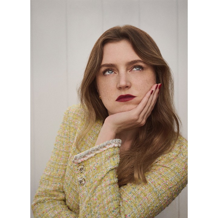 Portrait of a woman wearing a yellow tweed jacket with her hand supporting her chin against a plain vertical paneled wall