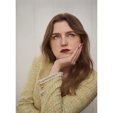 Portrait of a woman wearing a yellow tweed jacket with her hand supporting her chin against a plain vertical paneled wall
