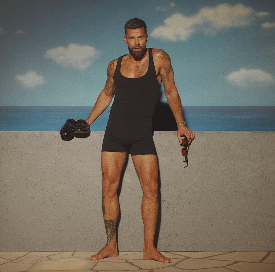 Man in vintage bathing suit stands barefoot against a faux beach background, holding shoes and sunglasses, looking into the camera.