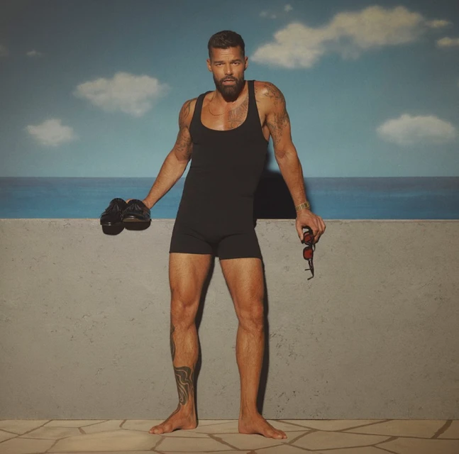 Man in vintage bathing suit stands barefoot against a faux beach background, holding shoes and sunglasses, looking into the camera.