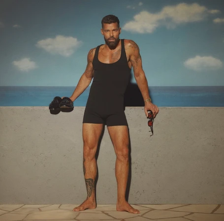 Man in vintage bathing suit stands barefoot against a faux beach background, holding shoes and sunglasses, looking into the camera.