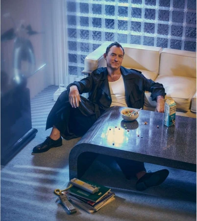 Male model in casual attire sits at a table with cereal, milk, and books in an indoor setting with a textured glass wall.