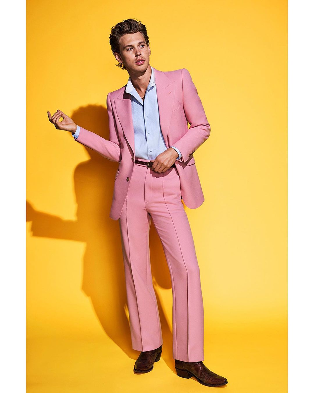 Portrait of a blonde adult male wearing a pink suit and brown western boots, standing against a yellow background in a studio.