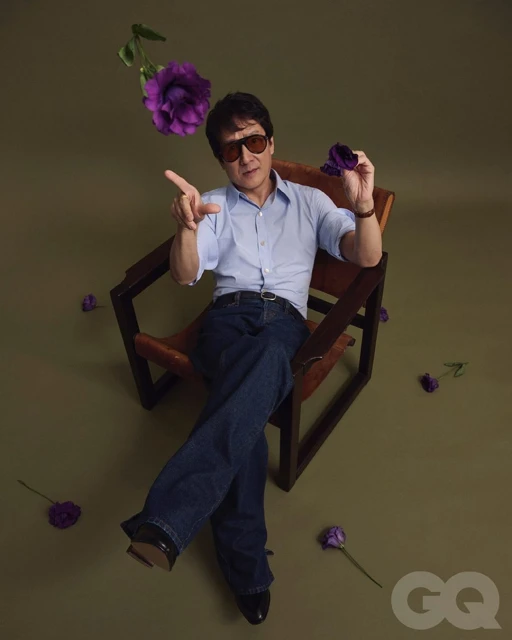 Adult male wearing jeans, light blue shirt, and sunglasses sits in a chair surrounded by purple flowers against a green background.
