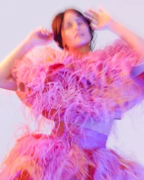 Model wearing a vibrant pink feathery textured garment with arms raised in a softly blurred, multi-colored neon-lit portrait.