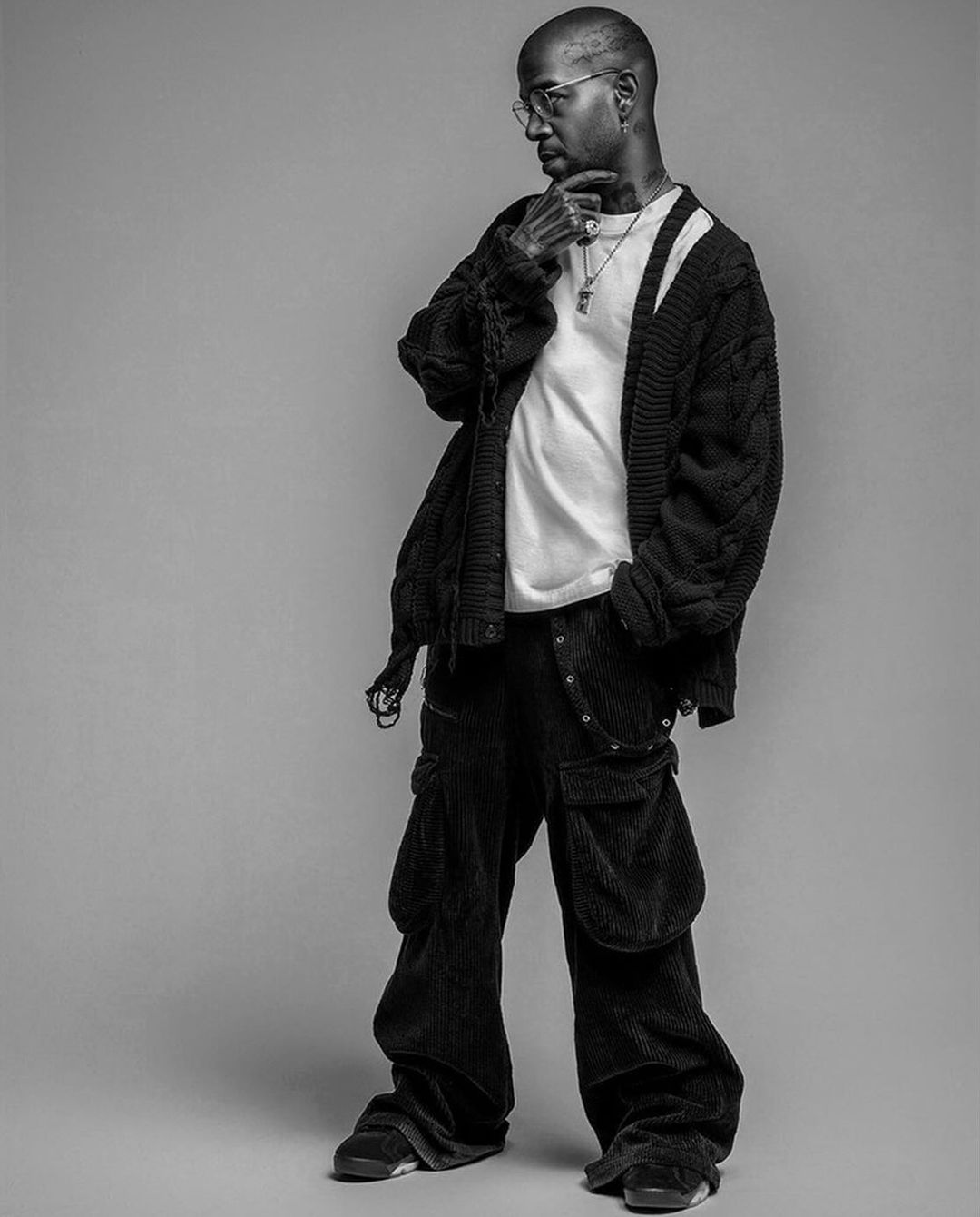 Monochromatic portrait of a stylish adult male in a casual white t-shirt, oversized cardigan, baggy pants, jewelry, and glasses looking left.