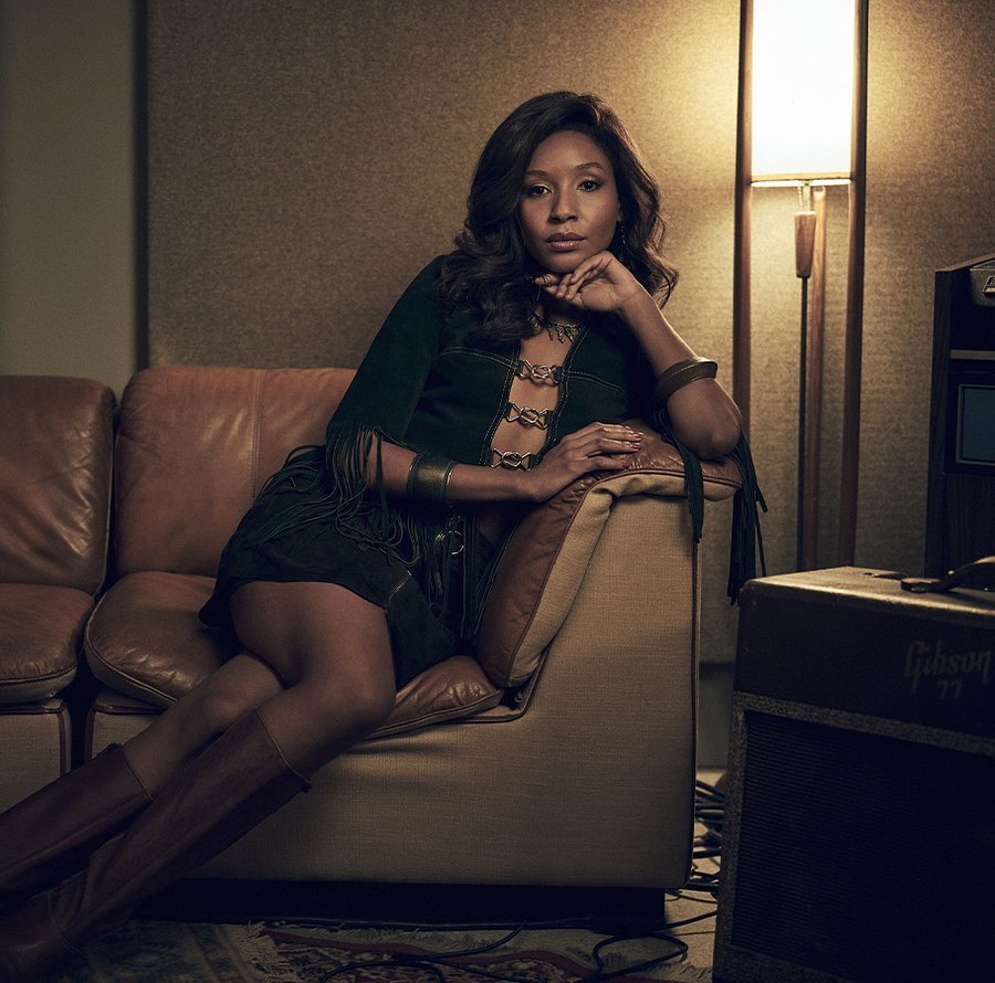 Woman with long brown hair sits on a brown leather couch in a studio wearing a suede green dress and brown boots.