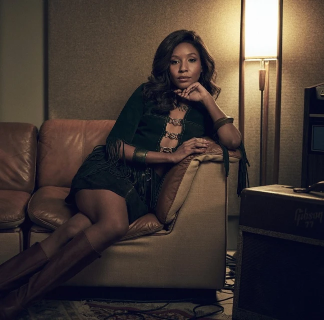 Woman with long brown hair sits on a brown leather couch in a studio wearing a suede green dress and brown boots.