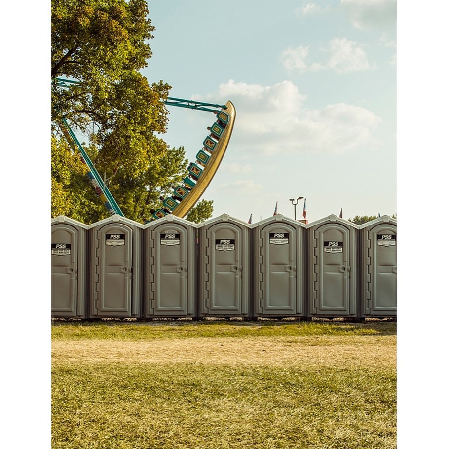 Row of portable toilets at an outdoor event, with green grass and a blurred carnival ride in the background under a blue sky.