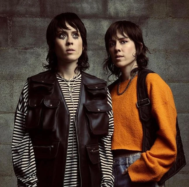 Two adult females stand indoors against a textured wall. One wears a striped shirt with a dark vest and the other, an orange sweater.