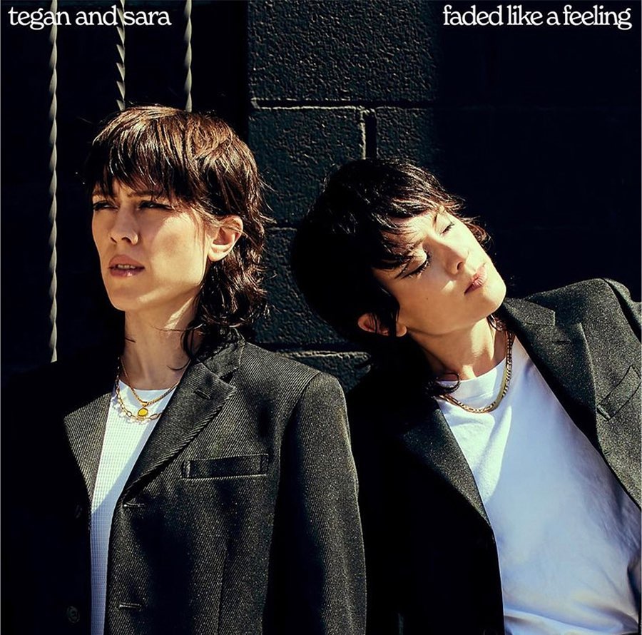 Two women with dark hair wearing black blazers and white t-shirts against a dark textured wall outdoors in sunlight.