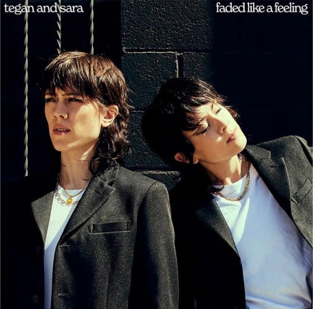 Two women with dark hair wearing black blazers and white t-shirts against a dark textured wall outdoors in sunlight.