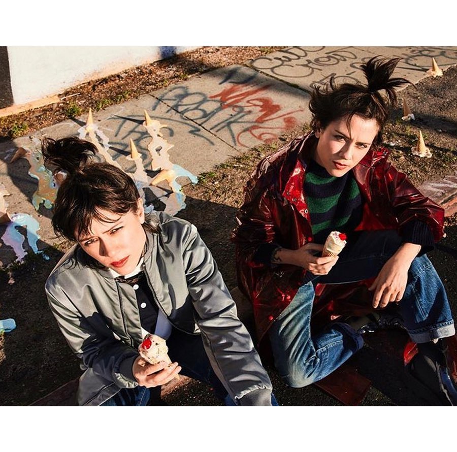 Two women sitting on concrete with melting ice cream cones, wearing jackets and jeans, captured in an eye-level outdoor shot.