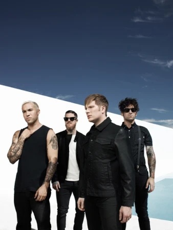 Four men in black clothing and sunglasses posing outdoors against a white architectural wall and blue sky.