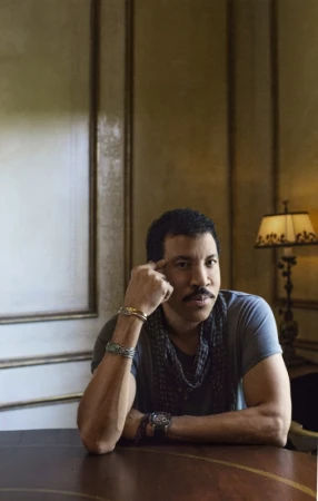 Lionel Richie seated indoors resting a hand on his temple wearing a grey t-shirt and layered bracelets photographed by Pamela Littky