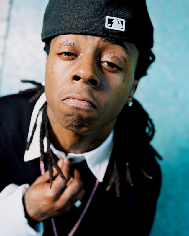 Close-up portrait of rapper Lil Wayne with dreadlocks, tattoos above his eyes, wearing a baseball cap indoors against a turquoise background.