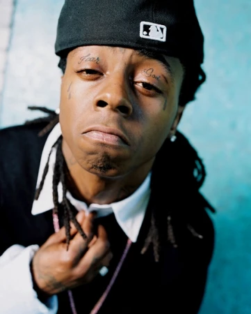 Close-up portrait of rapper Lil Wayne with dreadlocks, tattoos above his eyes, wearing a baseball cap indoors against a turquoise background.