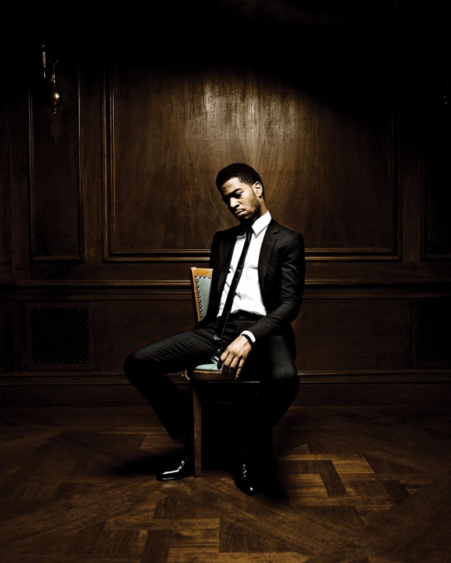 Adult male in a suit sitting on a chair indoors, with a wood panel wall background in dark lighting and a serious facial expression.