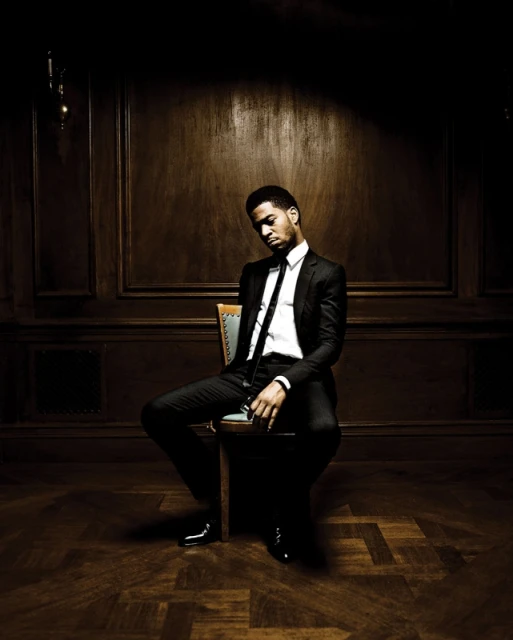 Adult male in a suit sitting on a chair indoors, with a wood panel wall background in dark lighting and a serious facial expression.