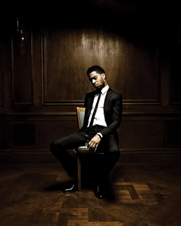 Adult male in a suit sitting on a chair indoors, with a wood panel wall background in dark lighting and a serious facial expression.