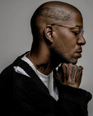 Profile portrait of Kid Cudi with facial and hand tattoos wearing glasses and a black knit sweater against a grey background.