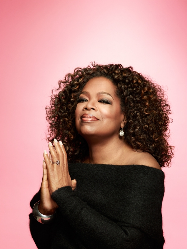 Portrait of Oprah Winfrey with hands folded, looking serene, against a pink background. She is wearing a black off-the-shoulder sweater.