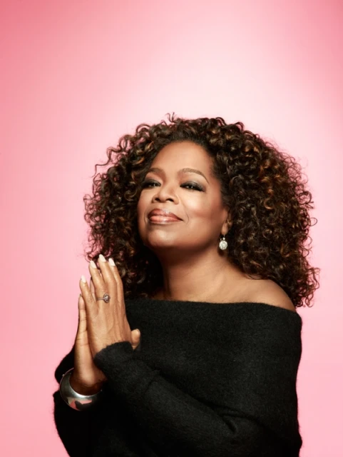 Portrait of Oprah Winfrey with hands folded, looking serene, against a pink background. She is wearing a black off-the-shoulder sweater.