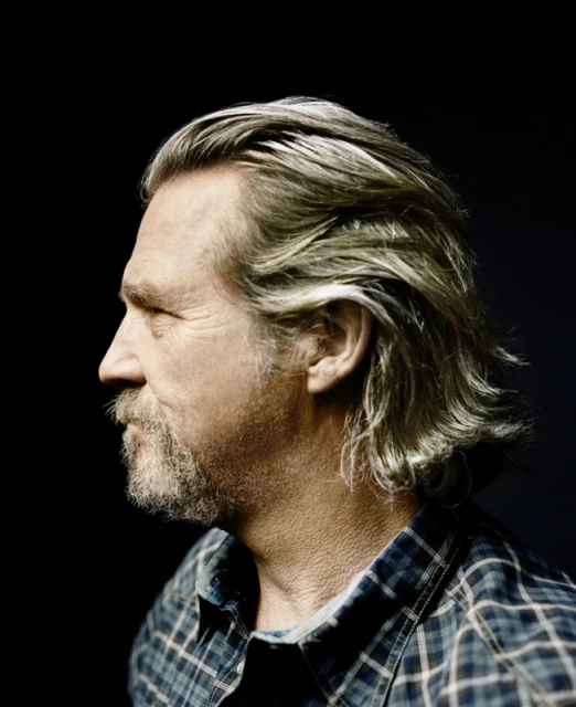 Profile portrait of an adult man with gray hair and a beard wearing a plaid shirt against a dark backdrop. Studio lighting highlights his features.
