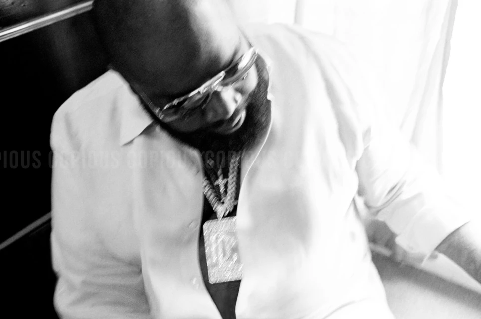 Black and white shot of a man with sunglasses, a beard, and a large chain in a light-colored shirt looking down.