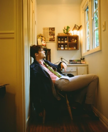 Man in dark coat and patterned shirt seated in narrow office space by a window, photographed by Pamela Littky