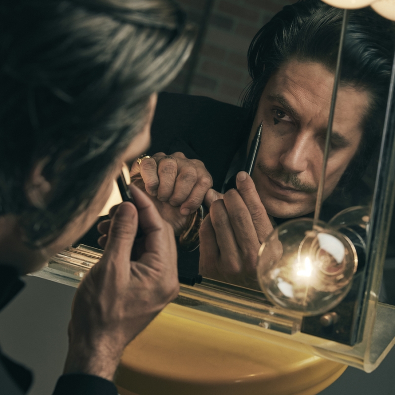 Adult male applying dark makeup in the mirror. A modern light fixture and beige wall are in the photo studio background.