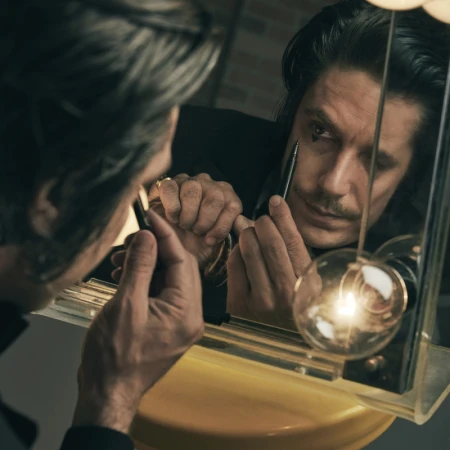 Adult male applying dark makeup in the mirror. A modern light fixture and beige wall are in the photo studio background.