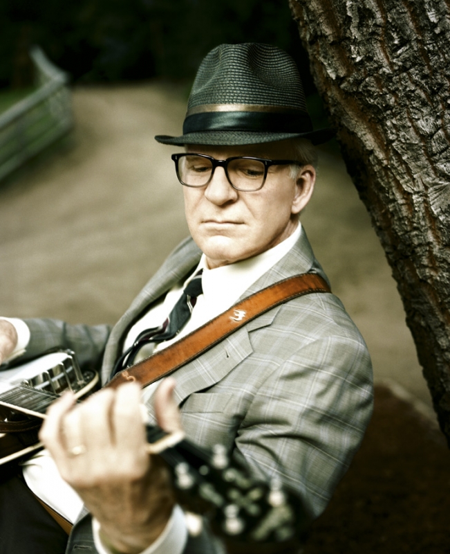 Portrait of a mature man in a fedora and glasses playing a banjo outdoors, leaning against a tree in a grey plaid suit.