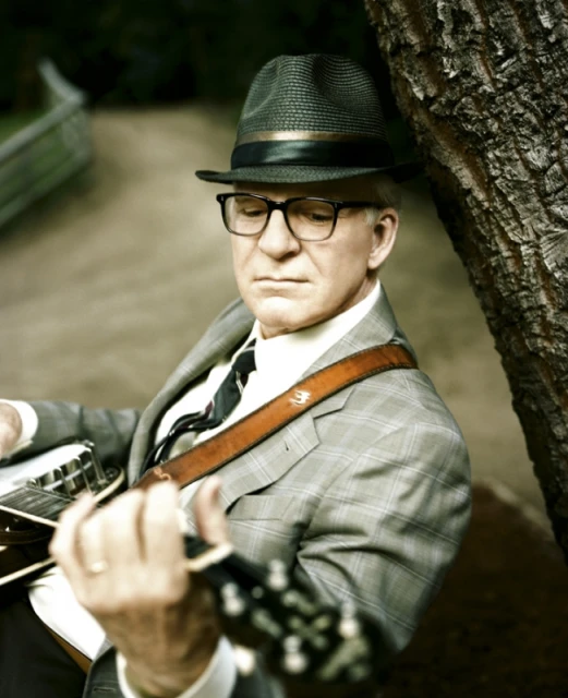 Portrait of a mature man in a fedora and glasses playing a banjo outdoors, leaning against a tree in a grey plaid suit.