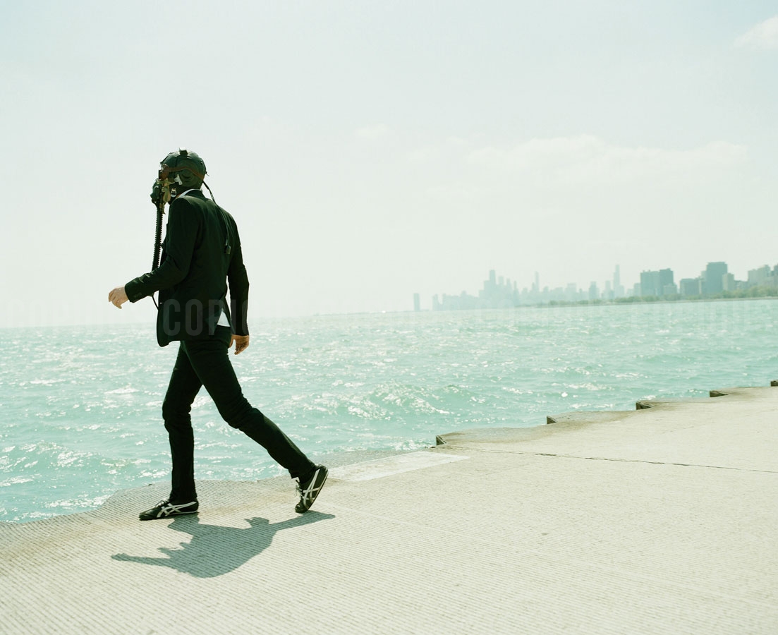 Adult male walking on pier in black suit and mask, facing the city skyline over the water on a hazy day. High angle, natural lighting.