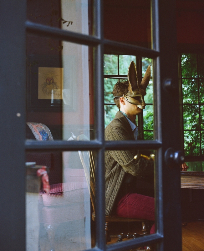 Person wearing a rabbit mask sits indoors, framed by glass doors. Red trousers, cardigan, and a view of leafy green trees.