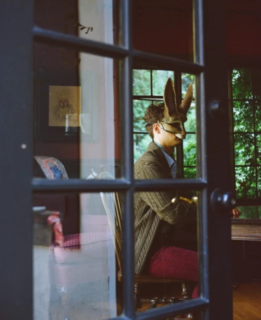 Person wearing a rabbit mask sits indoors, framed by glass doors. Red trousers, cardigan, and a view of leafy green trees.