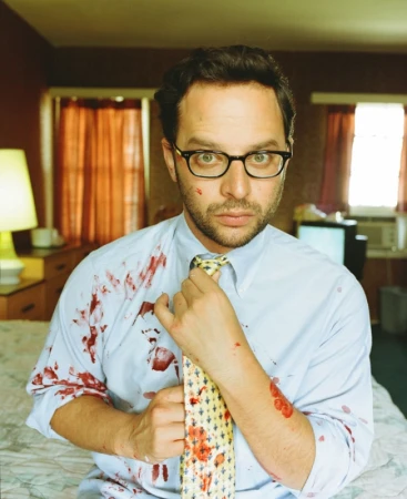Adult male with glasses in a light blue button-down shirt and patterned tie with blood spatter on his shirt, face and hands.