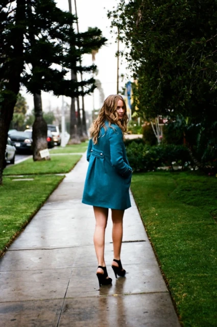 A woman in a teal coat and black heels walks away on a sidewalk lined with grass and trees under natural lighting conditions.
