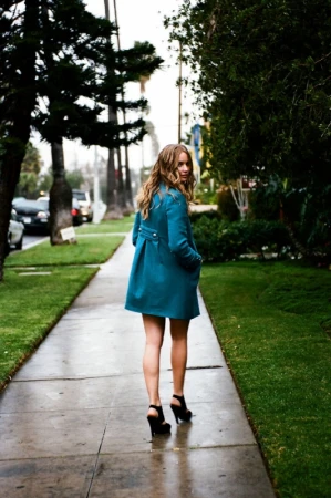 A woman in a teal coat and black heels walks away on a sidewalk lined with grass and trees under natural lighting conditions.