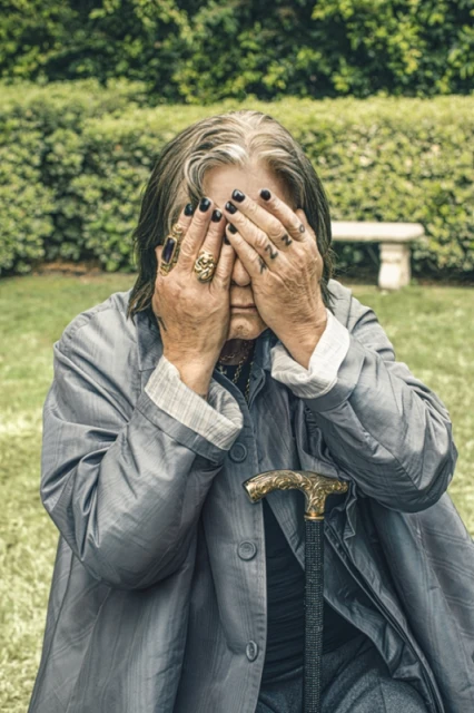 An elderly adult with long grey hair sits on green grass in front of manicured bushes, covering their face with their hands.
