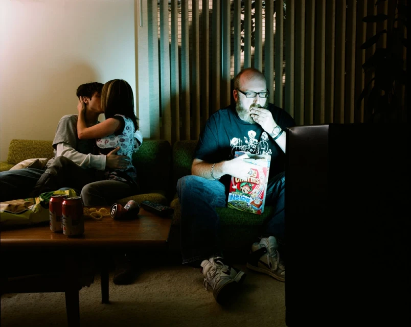 A couple kissing on a green couch while a bearded man eats cereal in front of a television inside a dimly lit room.