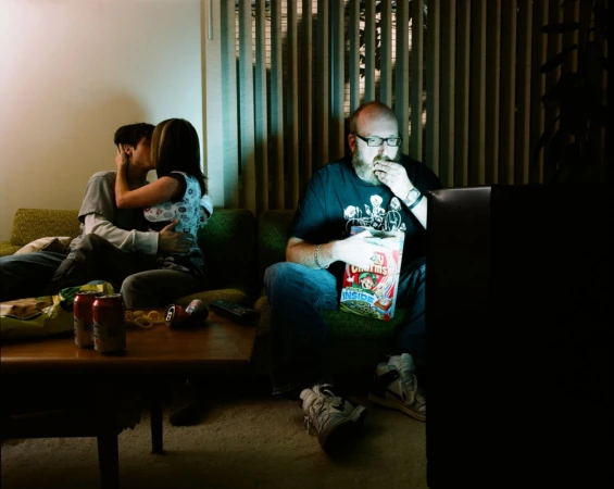 A couple kissing on a green couch while a bearded man eats cereal in front of a television inside a dimly lit room.