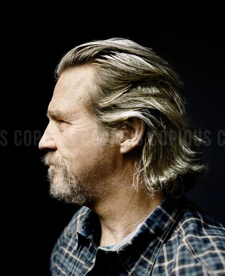 Profile portrait of Jeff Bridges with long grey hair and a beard, wearing a dark plaid shirt against a dark background.