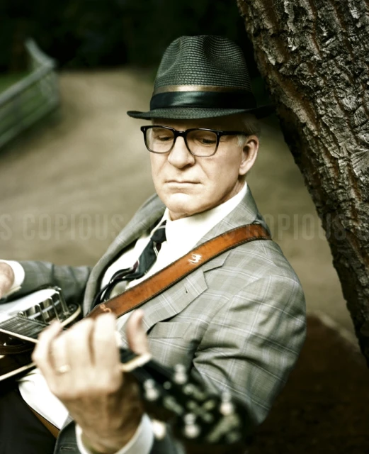 Adult man plays banjo outdoors: He wears a hat, glasses, suit, and tie, leaning against a tree on a path in a park-like setting.