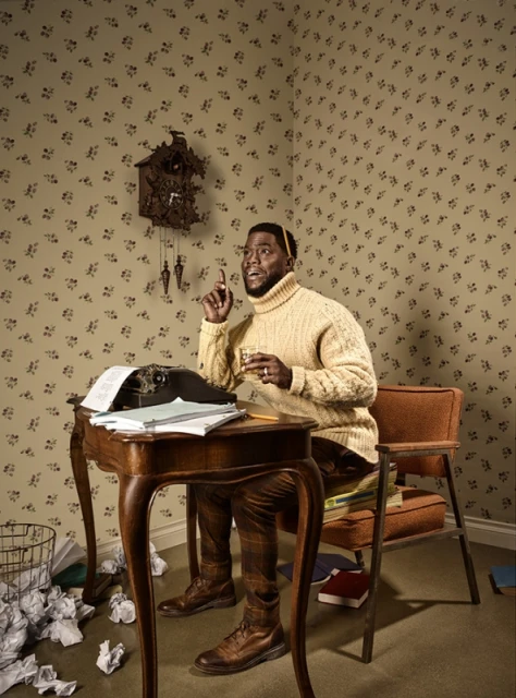 Kevin Hart sits at a wooden desk with a typewriter in a room with patterned wallpaper, wearing a yellow sweater and brown plaid pants.