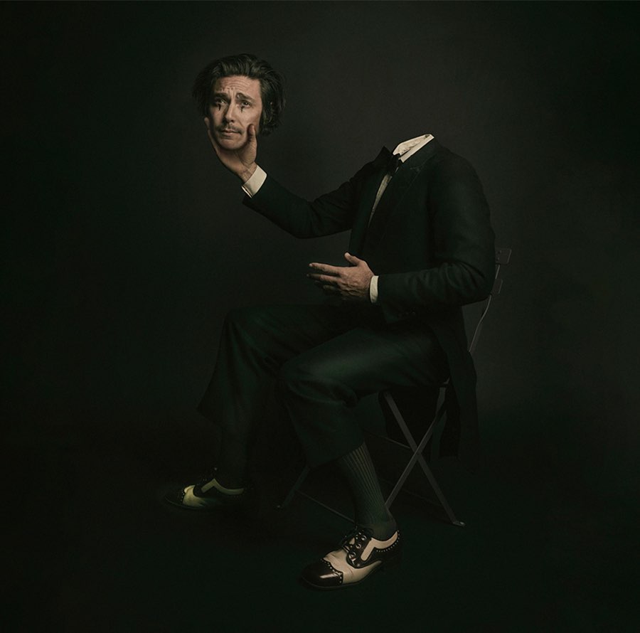 A man in a suit sits with his head levitating in his hand against a dark background. Studio lighting creates a surreal portrait.
