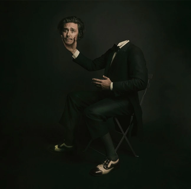 A man in a suit sits with his head levitating in his hand against a dark background. Studio lighting creates a surreal portrait.