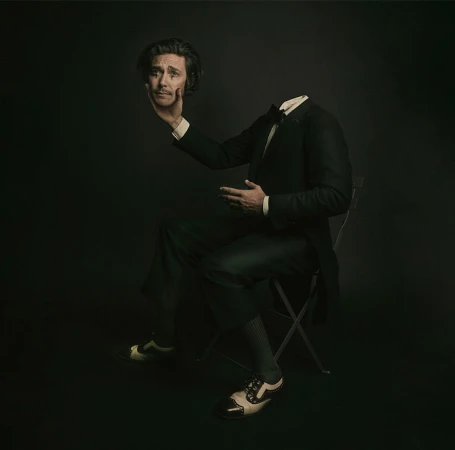 A man in a suit sits with his head levitating in his hand against a dark background. Studio lighting creates a surreal portrait.
